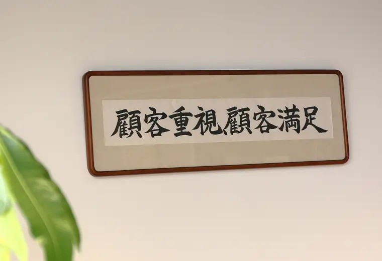 壁に掛けられた額縁入りの書作品。「顧客重視顧客満足」の8文字が墨で書かれ、左下に観葉植物の葉が写る