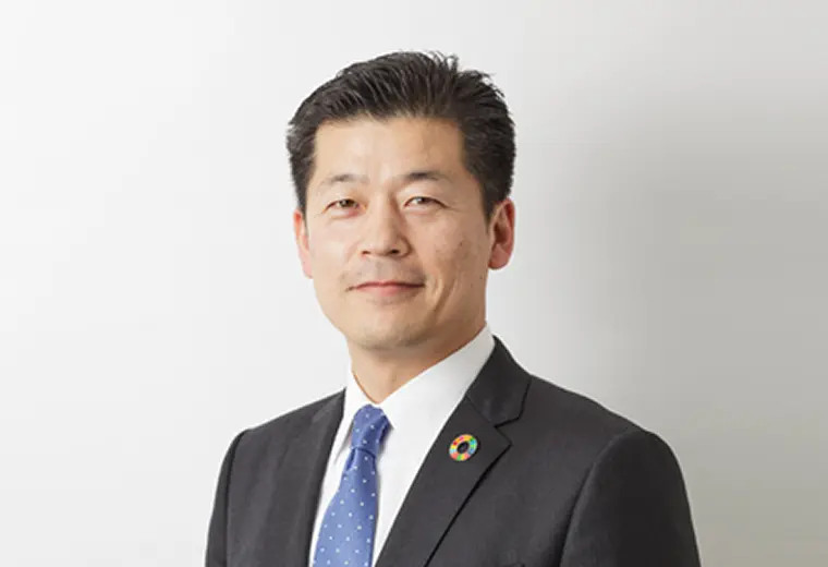 薄いグレー背景のビジネスポートレート。ダークスーツと水色のドット柄ネクタイを着用し、左襟にSDGsのカラフルなバッジを付けた男性が正面を向いて微笑む