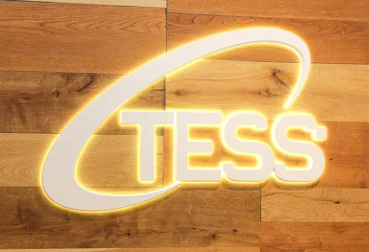 横並びの木目壁に設置された「TESS」の白い立体ロゴ。文字と上部の弧状ラインが黄色い逆光で光る