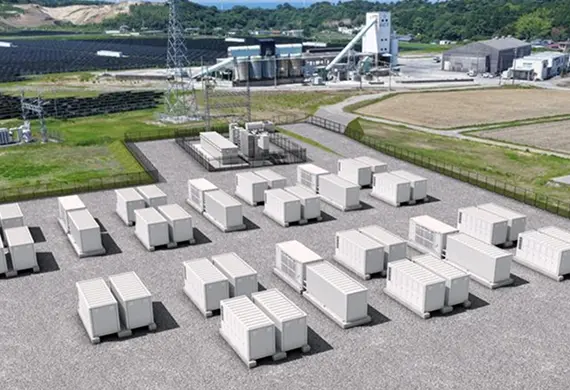 上空から見た蓄電施設。白いコンテナ型バッテリーが並ぶ敷地、送変電設備と鉄塔、背後の太陽光発電所と工場、丘陵地を背景にしたエネルギー拠点