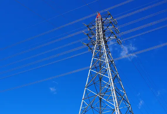 青空を背景に見上げた鋼製の送電用鉄塔。碍子と送電線、塔頂の赤い部品が写る
