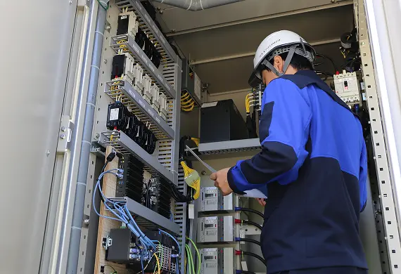 開いた制御盤の前で、白いヘルメットと作業着の作業員が書類を手に配線やブレーカーを点検する様子