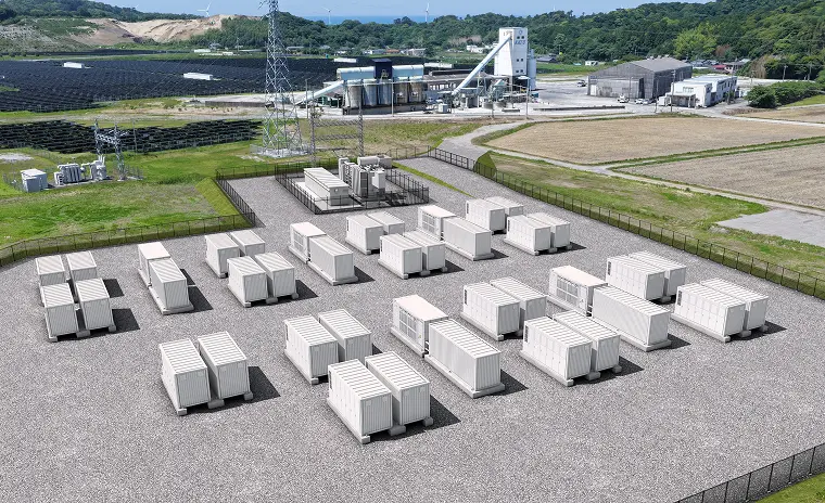 上空から見た蓄電施設。フェンス内に白いコンテナ型バッテリーが並び、左に太陽光発電所と送電鉄塔、奥に工場と倉庫、丘陵と海、遠方に風力発電機