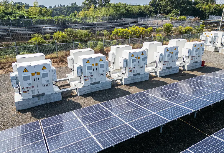 傾けた太陽光パネル手前に、砂利地で七基以上並ぶ白いパワコン筐体。高圧注意表示、ケーブルトレー、フェンス向こうの緑の丘陵