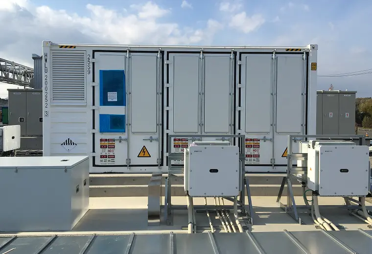 コンクリート上の白いコンテナ型蓄電設備。手前に二基のパワコン筐体とケーブル、扉に高電圧・危険の警告表示と容器番号、青空の下