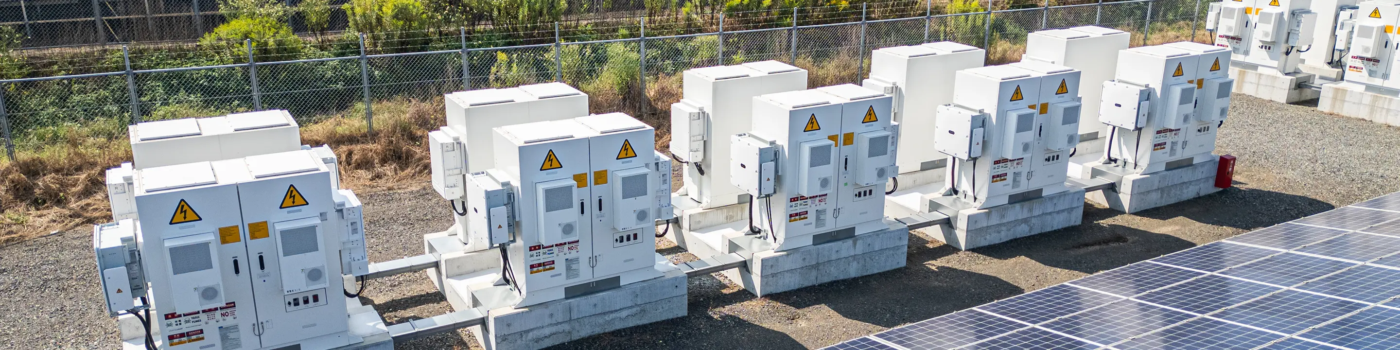 太陽光発電所の砂利地に並ぶ白いパワコン筐体の全景。各機に高圧注意表示、手前右にパネル列、ケーブルトレーで連結、奥にフェンスと緑の樹木