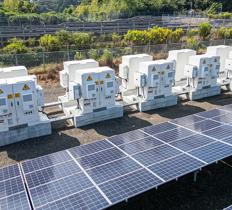手前に傾けた太陽光パネルの奥に、コンクリート基礎上で五基並ぶ白いパワコン筐体。高圧注意表示と地上ケーブルトレー、フェンス向こうのパネル列と緑の丘陵