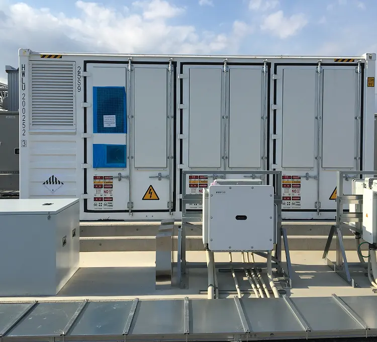 屋外の白いコンテナ型蓄電設備。手前に蓄電池ブロックと書かれた制御ボックス、高圧・毒性煙の警告ラベル、背景に送電設備と緑の丘陵