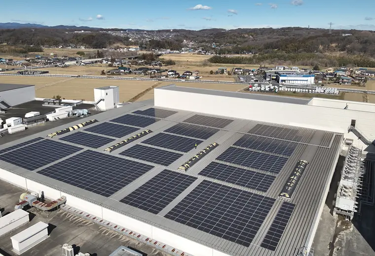 大規模工場の平屋根に区画ごとに敷かれた太陽光パネル群の空撮。黄色い安全ベストの作業員が屋上通路に1人立ち、周囲に茶色の畑や住宅、遠くの丘陵が広がる