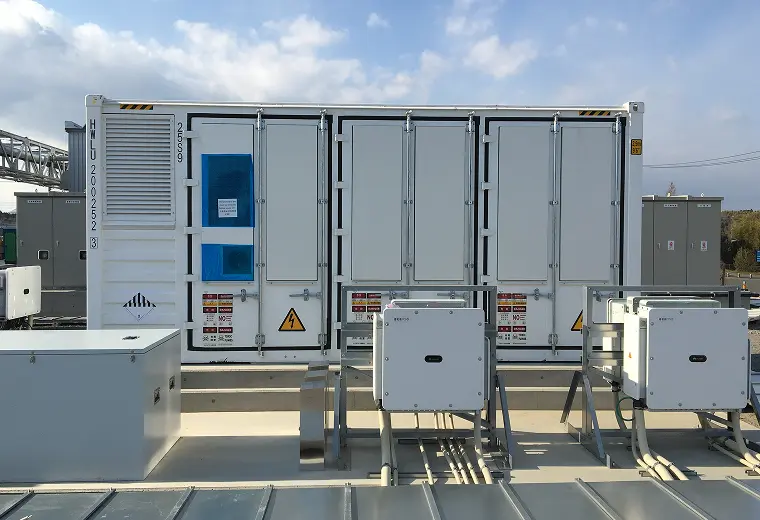 屋外のコンクリート基礎に設置された白いコンテナ型蓄電設備。手前にHuaweiロゴのパワーコンディショナ2台と配線、コンテナ扉には高電圧などの警告表示