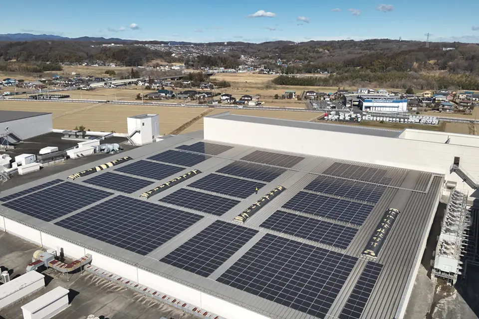 大規模工場の平屋根に区画ごとに敷かれた太陽光パネル群の空撮。黄色い安全ベストの作業員が屋上通路に1人立ち、周囲に茶色の畑や住宅、遠くの丘陵が広がる