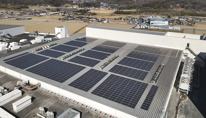 工場の平屋根に区画ごとに並べられた太陽光パネル群の空撮。黄色い安全ベストの作業員が通路に1人立ち、周辺に住宅地や田畑、遠くに山並みが広がる