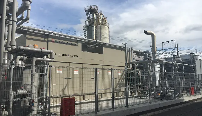 チェーンリンクフェンス越しに見る三菱重工エンジン＆ターボ社の1,000kWガスエンジン発電装置。ベージュ色のカバーと周囲の配管、背後のサイロや排気設備