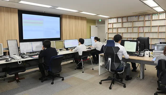 壁面の大型モニターと複数台のデスク、モニターを前に作業する複数人のスタッフ。右壁一面の分類帳（バインダー）の並んだ監視・運用オフィス