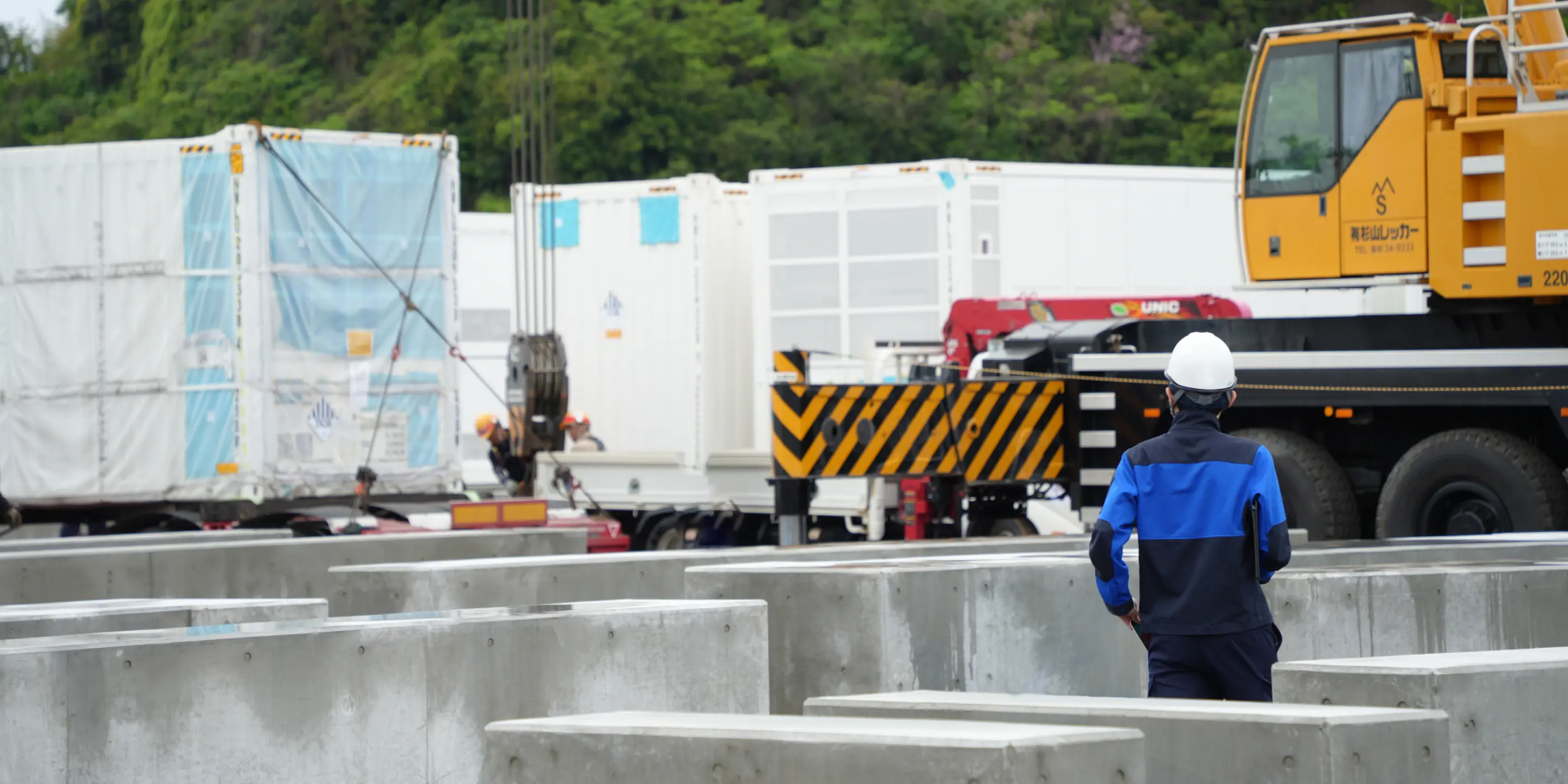 建設資材が並ぶ屋外ヤードで、クレーン車やコンテナを背にして作業を見守るヘルメット姿の作業員