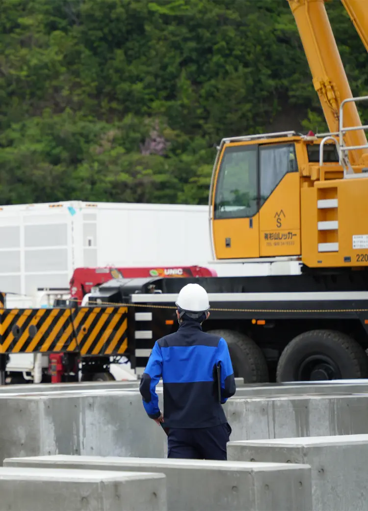 建設資材が並ぶ屋外ヤードで、クレーン車やコンテナを背にして作業を見守るヘルメット姿の作業員