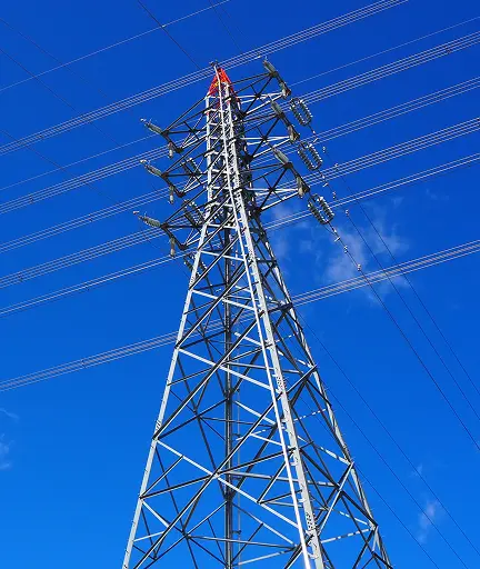 青空を背景に仰角で撮影した鋼製鉄塔の送電用鉄塔。碍子と数本の高圧送電線、塔頂の赤色部が写る